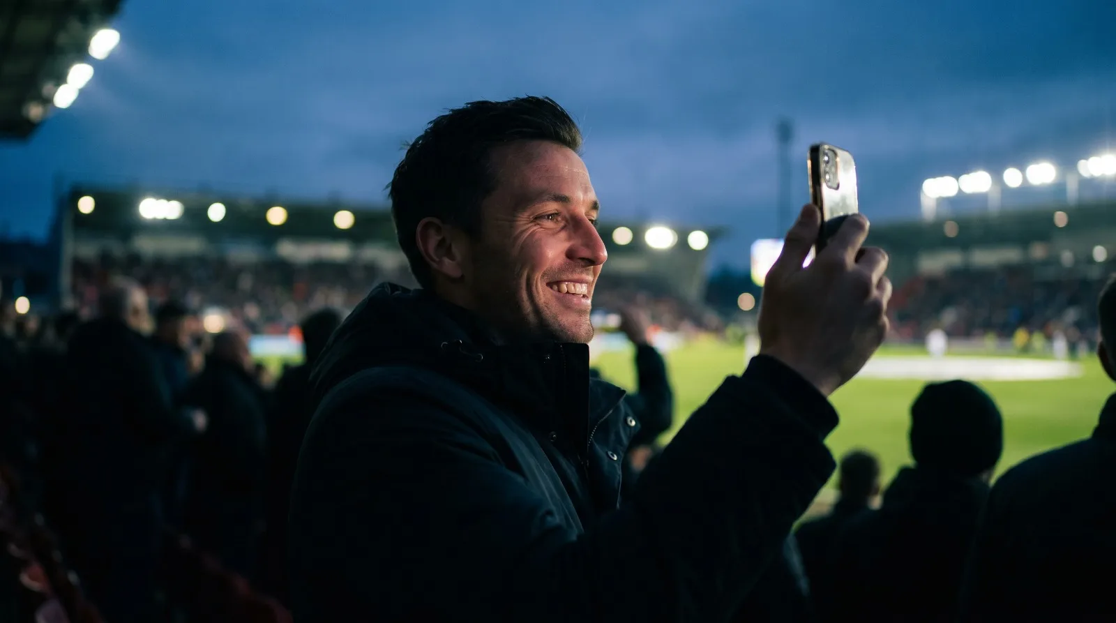 Um close-up de um homem sorridente de perfil em um estádio de futebol à noite. Ele segura um smartphone e usa um smartwatch de alta tecnologia, ambos emitindo um brilho gélido de ciano e azul, com gráficos de interface de apostas Ice.bet e padrões de flocos de neve digitais. Os refletores do estádio ao fundo têm um tom de azul frio, e outros torcedores desfocados completam a cena."